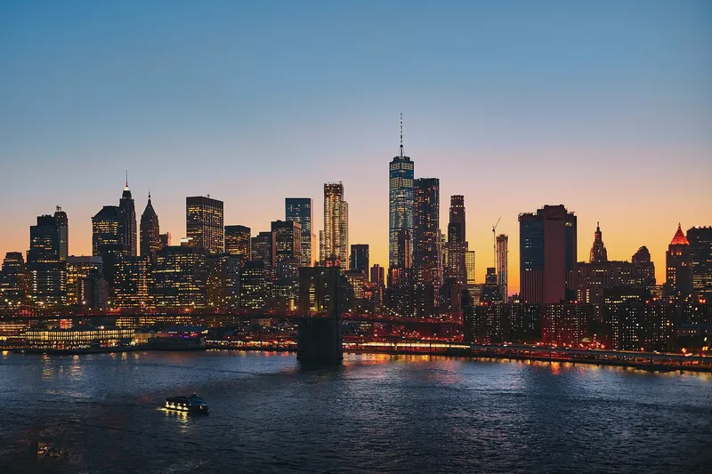 Lower Manhattan skyline at dusk with the Brooklyn Bridge crossing the East River in the foreground