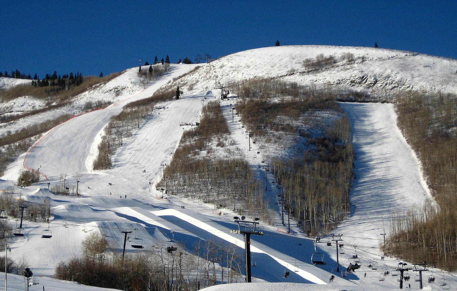 Skiers riding a chairlift above groomed slopes toward a snowy mountain summit on a clear winter day
